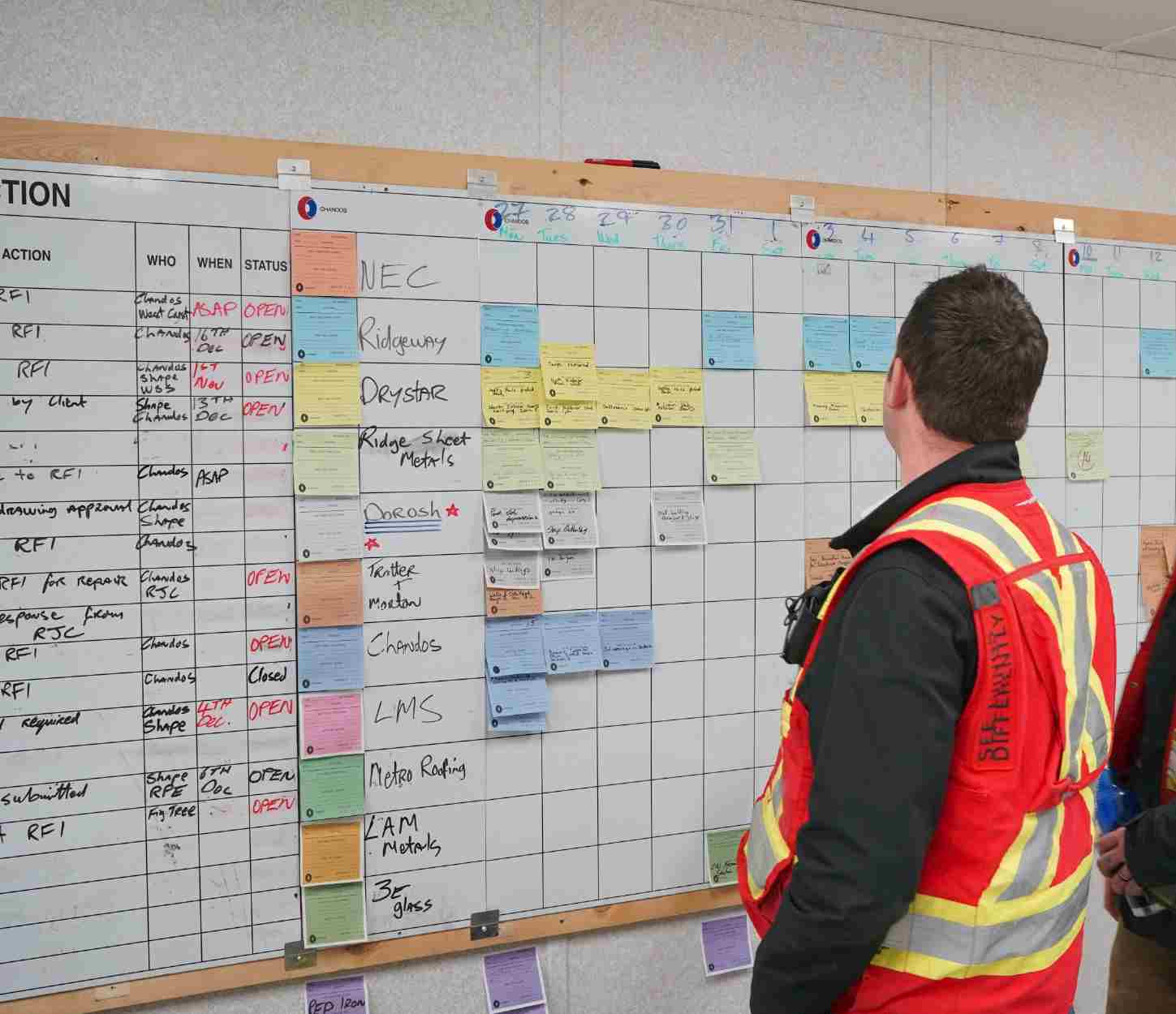 A construction professional in a safety vest stands in front of a large white board covered in colorful sticky notes representing tasks and schedules for a construction project. He appears to be reviewing the information on the board.